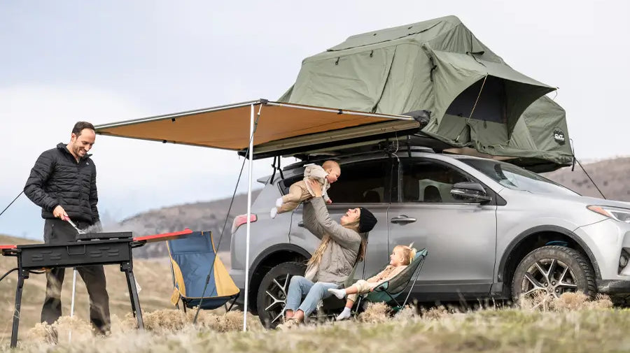 A family relaxing under an awning accessory attached to a rooftop tent above an SUV, while a man cooks on a grill.